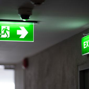 A green fire exit sign is placed on the ceiling along the dimly lit corridor and there is green exit sign on the exit door.